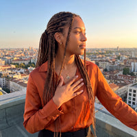 Thumbnail for Woman with long braids on a rooftop at sunset, hand on her heart, wearing the gold secret clover photo necklace | SKU: TPH-CLVN-GOLD