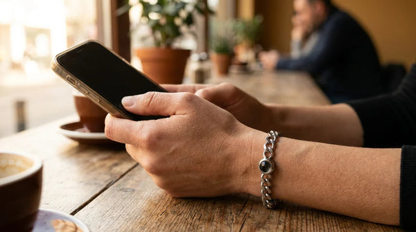 A woman holding a smartphone over her left wrist resting on a wooden cafe table, the Brother & Sisters Steel Photo Bracelet visible with its projection lens bead, illustrating how the hidden photo reveals through a phone camera