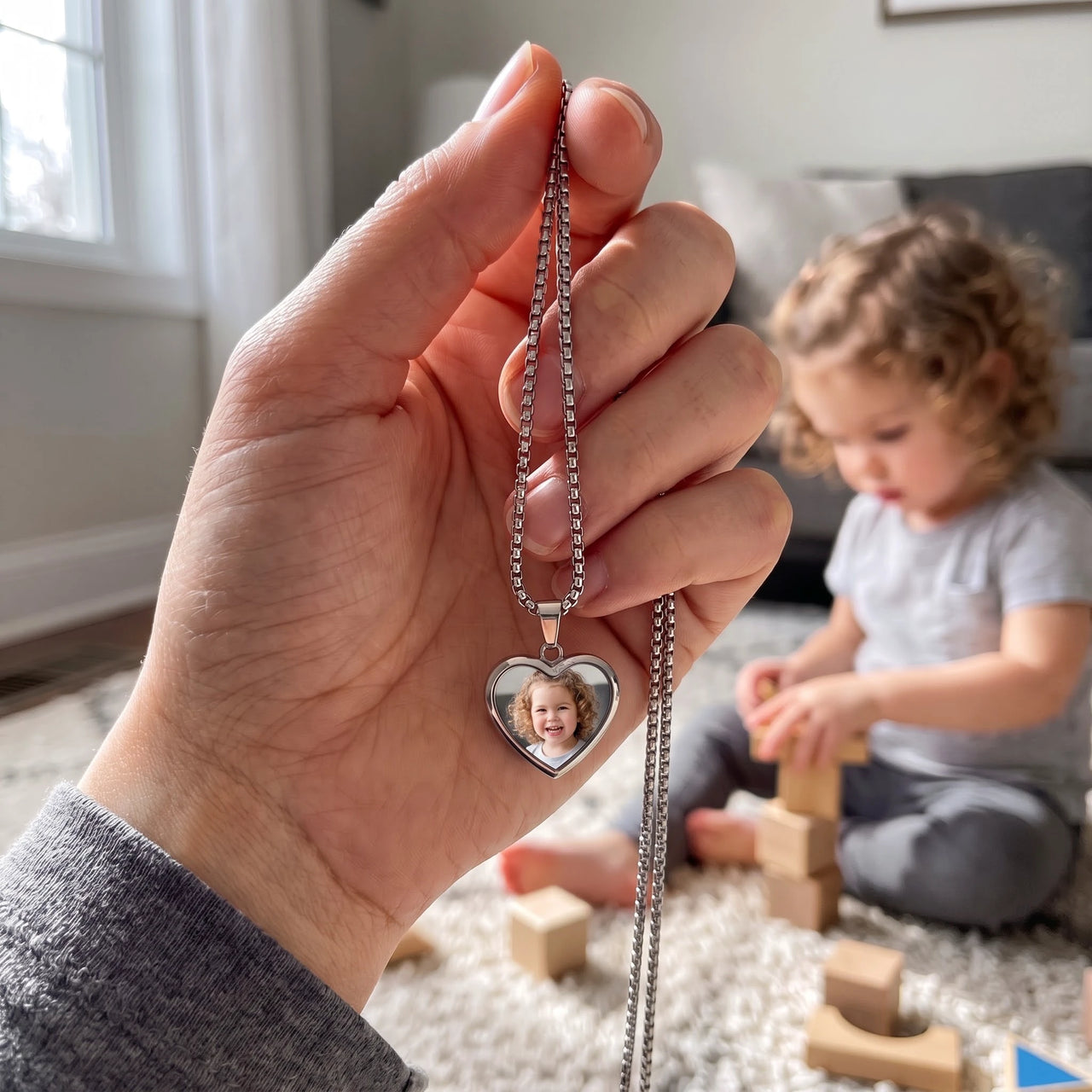 The silver heart photo pendant held up, real small size, her little girl playing behind | SKU: TPH-CHTN-SILVER-SMALL
